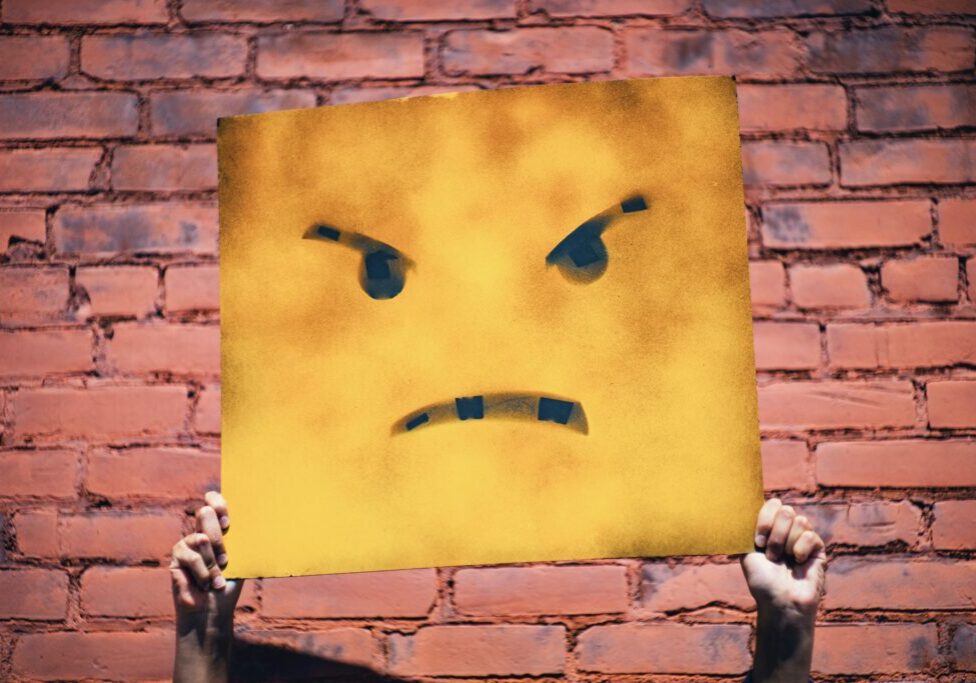 A person holding up a yellow sign with an angry face on it.